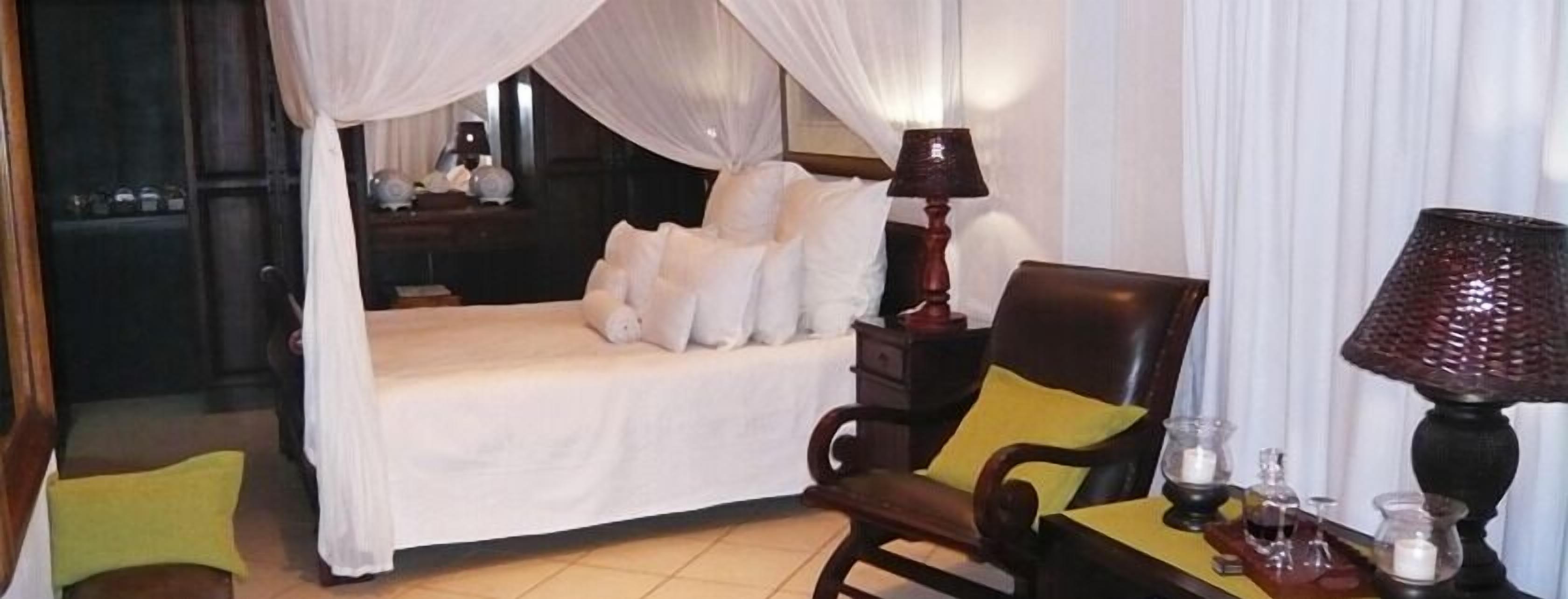 A bedroom with a four-poster bed, a chair with a yellow cushion, a bedside table with a lamp, and a mirror.