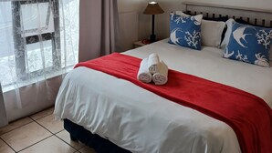 Habitación de hotel con un ventanal grande, cama con ropa blanca y detalles rojos, una lámpara y almohadas decorativas.