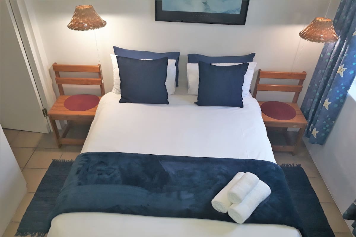 Ein ordentlich gemachtes Bett mit einer dunkelblauen Decke und Kissen, zwei hölzerne Stühle mit roten Sitzkissen und zwei Wandleuchten.