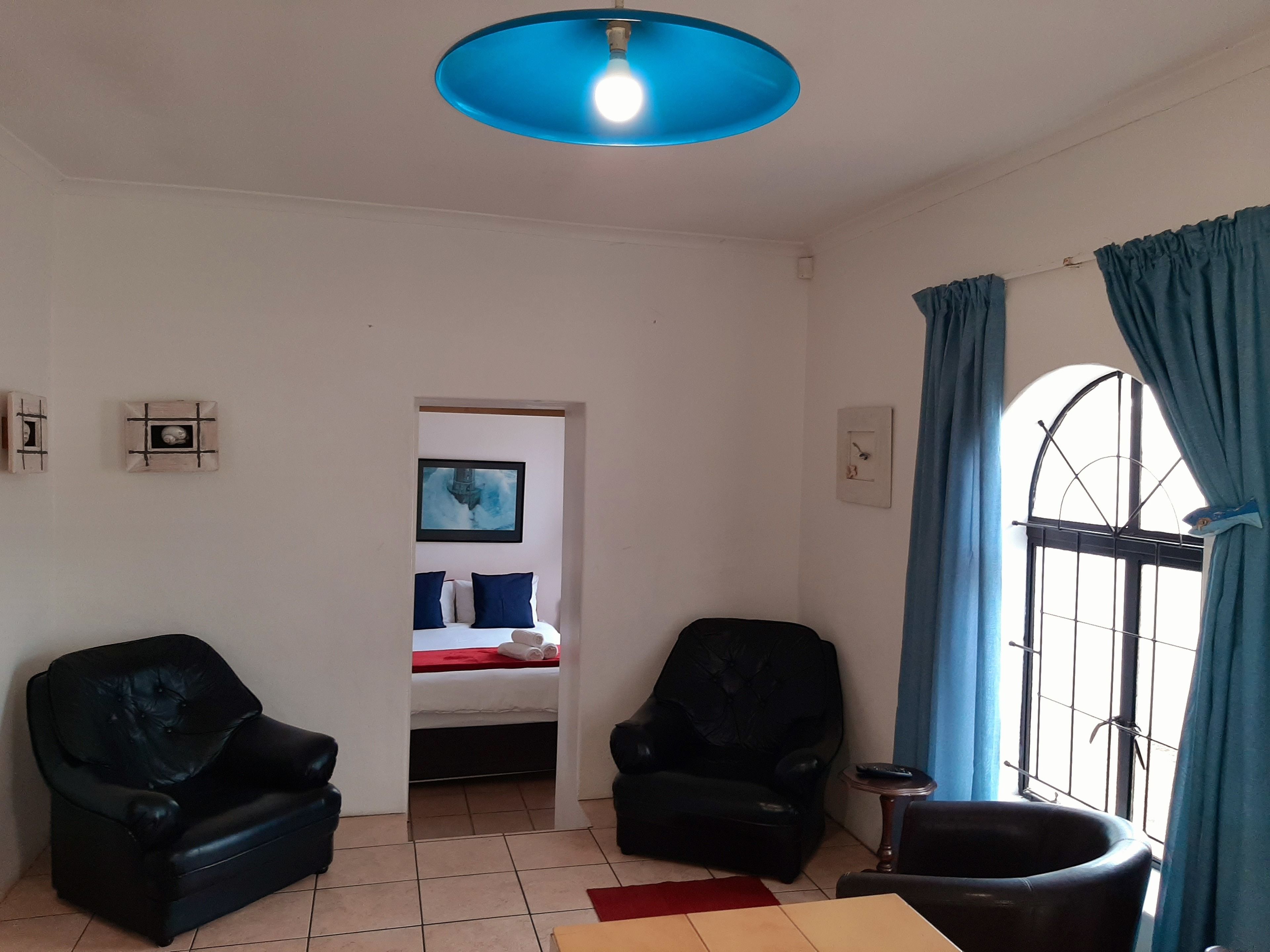 Una habitación con dos sillones de cuero negro, una cama con manta roja, una lámpara azul y una ventana con cortinas azules.
