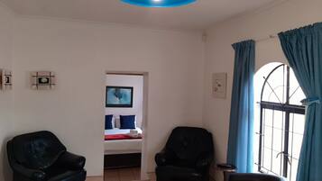 Una habitación con dos sillones de cuero negro, una cama con manta roja, una lámpara azul y una ventana con cortinas azules.