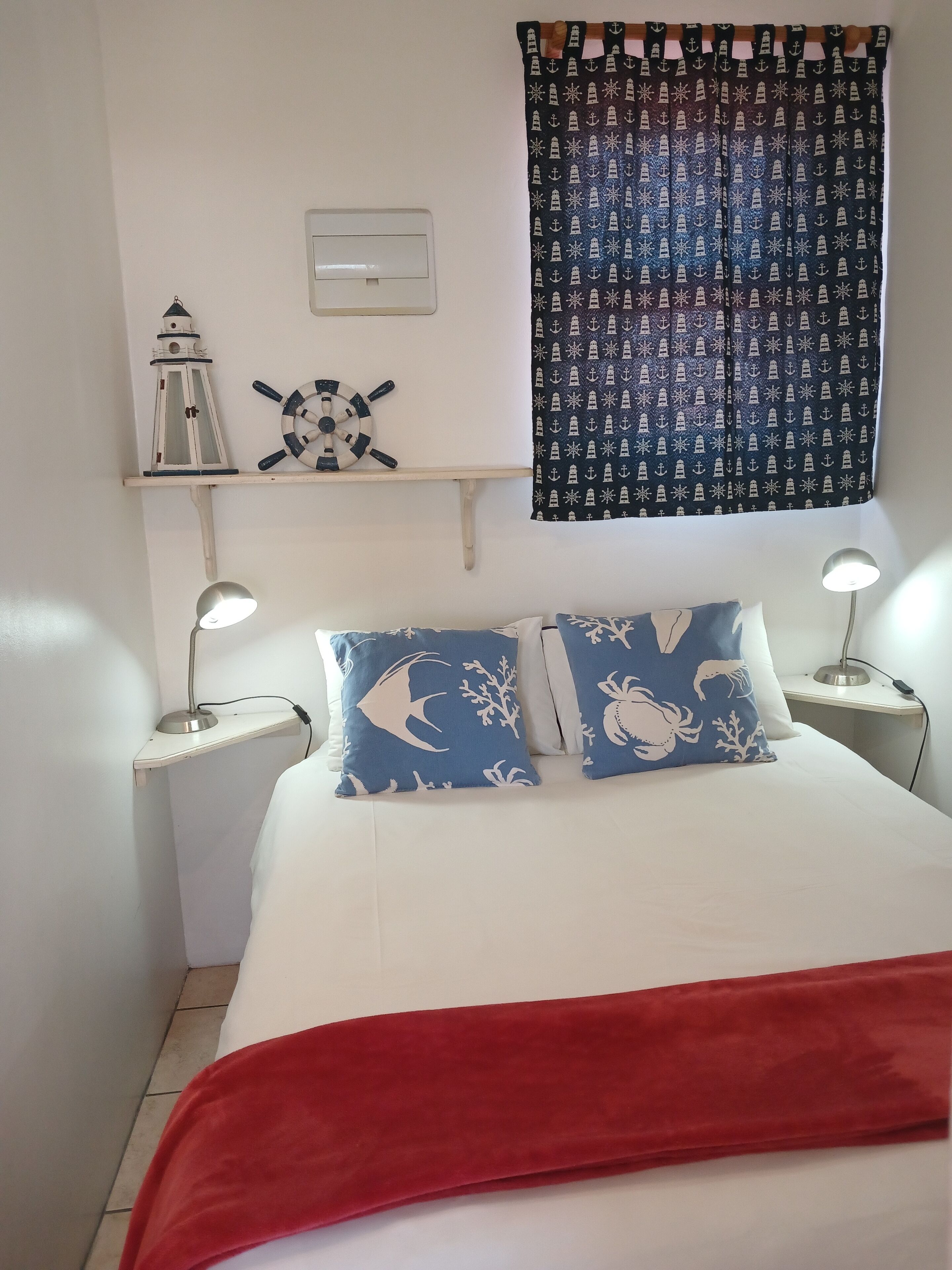 A bedroom with a bed, two nightstands, a wall-mounted shelf with a lantern and a ship's wheel, a patterned curtain, and a red blanket.