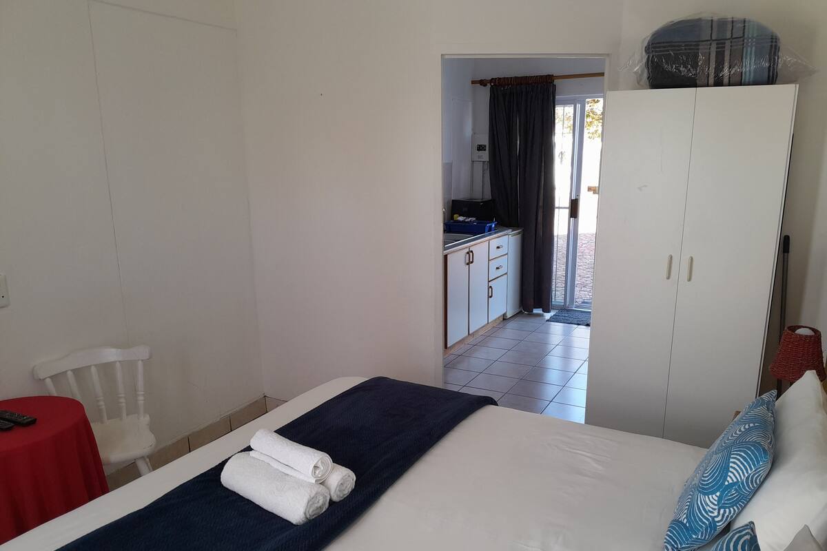 Ein Schlafzimmer mit einem Bett, einem roten Tisch, einem weißen Stuhl, einem Kleiderschrank und Blick auf eine Küche und einen Balkon.