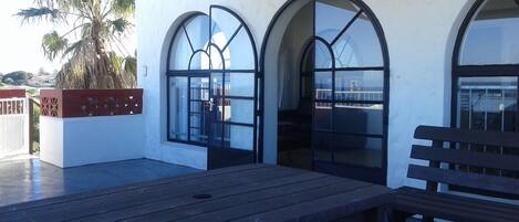 Un espace patio avec une table et un banc en bois, deux fenêtres en arc de cercle et un jardinier contenant un palmier.