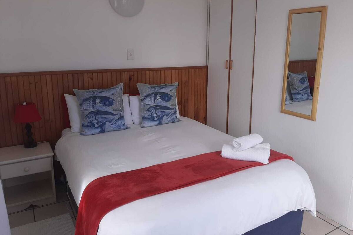 Ein ordentlich bezogenes Bett mit roter Decke, zwei Kissen mit blauem Fischmuster, ein Nachttisch mit roter Lampe und ein Spiegel an der Wand.