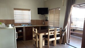 A compact kitchen with a dining table, chairs, a refrigerator, and a TV mounted on the wall. - Tourist Lodge Gansbaai (Gansbaai)