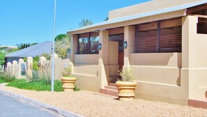 A single-story building with a flat roof, beige exterior walls, and a small porch area. There are two large potted plants on the porch. - Tourist Lodge Gansbaai (Gansbaai)