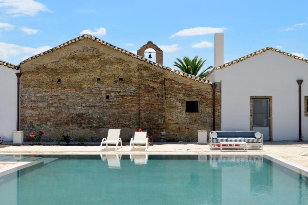 Un área de piscina con un edificio de piedra, dos sillones blancos y un sofá azul.