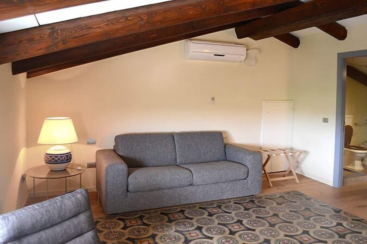 Ein Wohnzimmer mit grauem Sofa, einem gemusterten Teppich, einer Lampe und einem Holzbalken an der Decke.