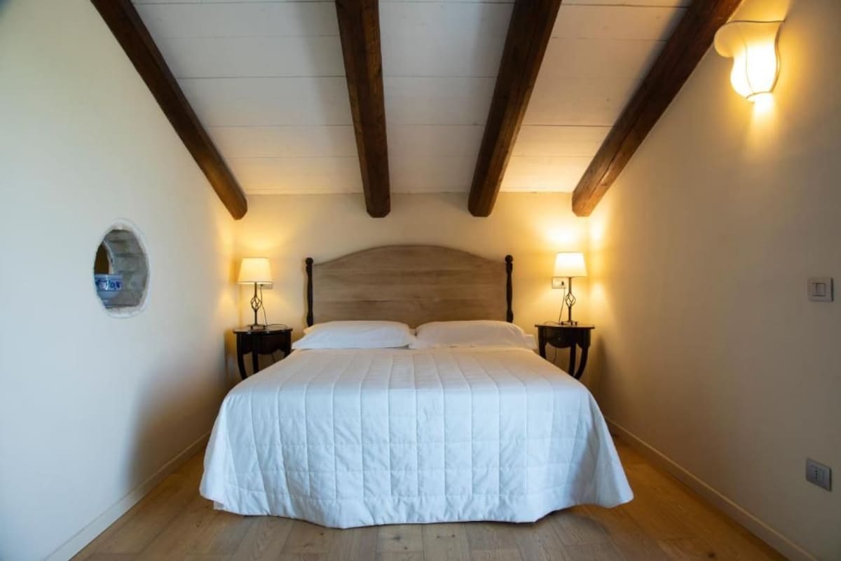 Un dormitorio con una cama grande, vigas de madera en el techo, dos lámparas de noche y una decoración de pared de piedra.