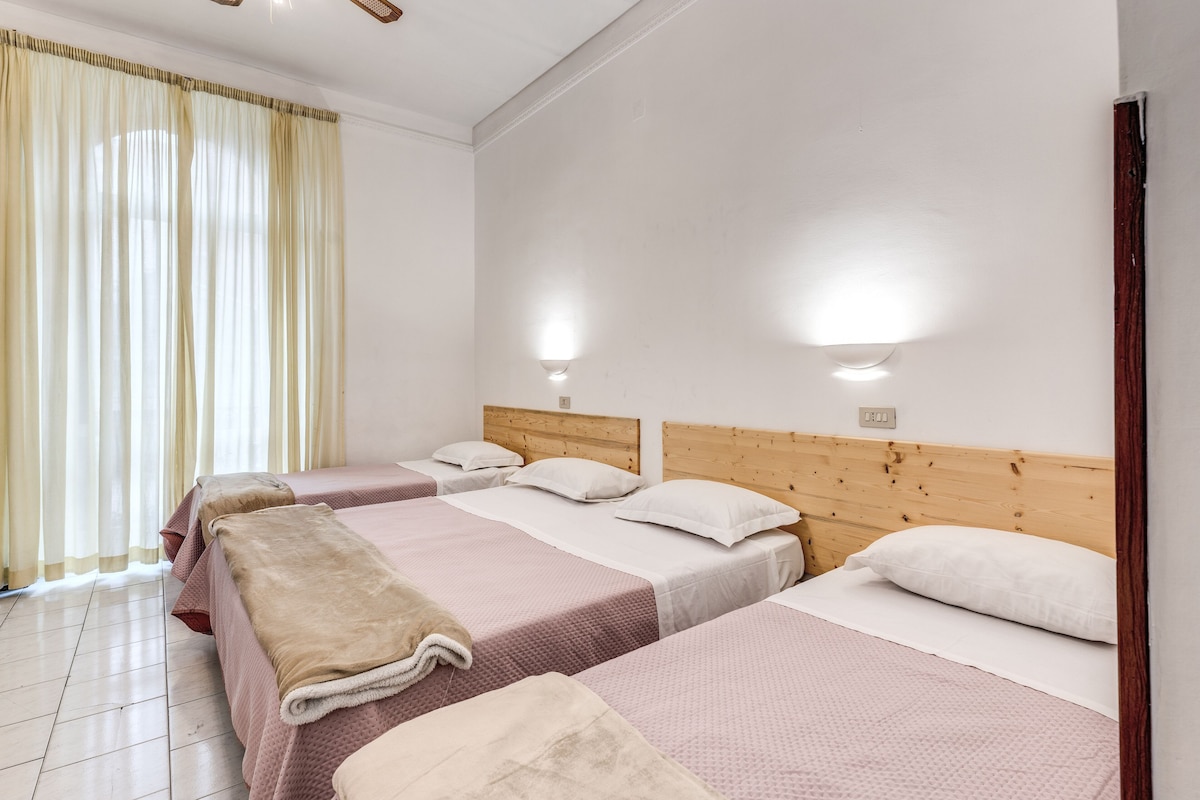 A hotel room with three beds, each with a wooden headboard, white bedding, and a beige throw blanket on one bed. There are two wall-mounted lights and a window with curtains.