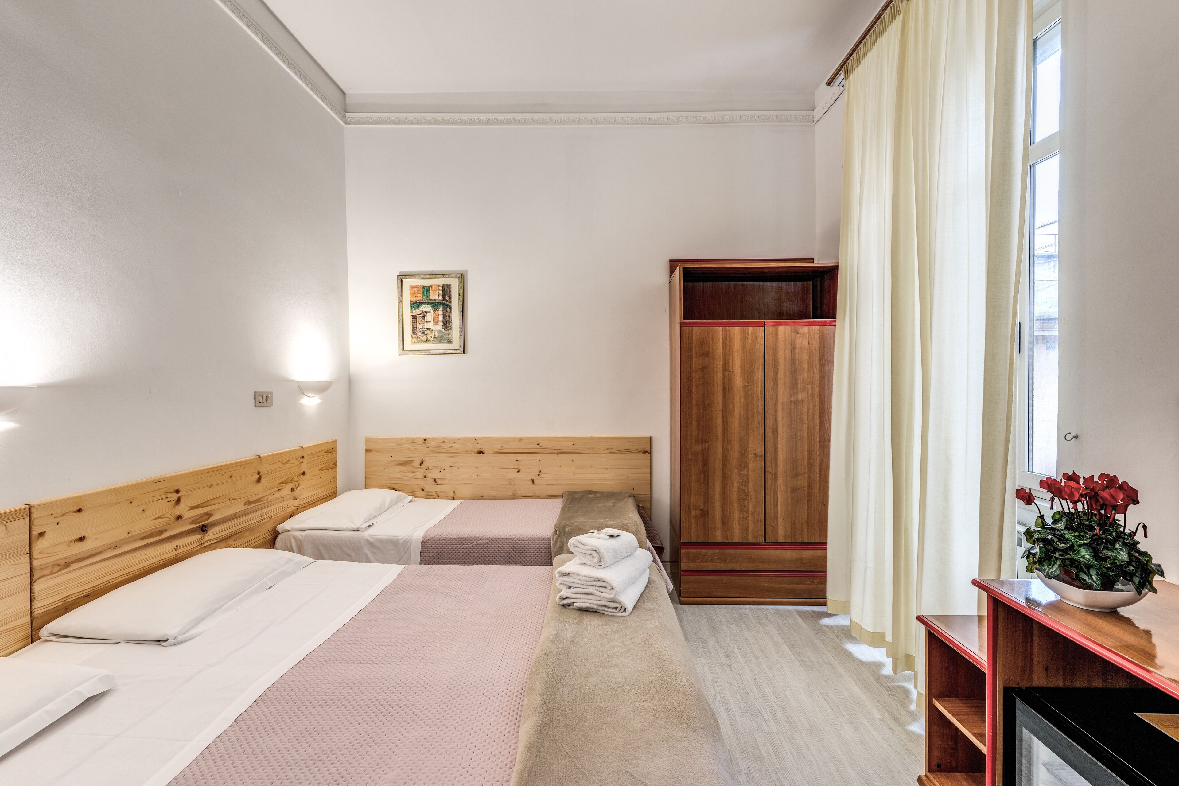 Une chambre d’hôtel avec deux lits, une armoire en bois, une table de chevet ornée d’un vase de fleurs et un tableau encadré au mur.