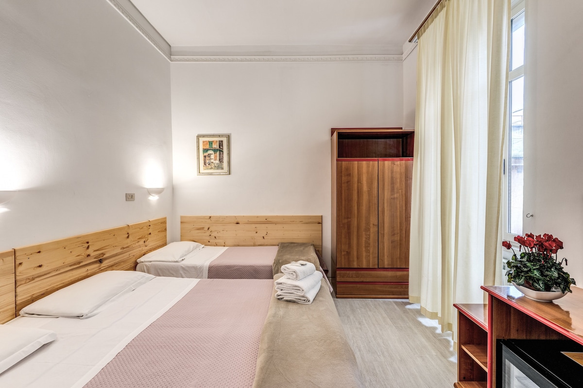A hotel room with two beds, a wooden wardrobe, a bedside table with a vase of flowers, and a framed picture on the wall.