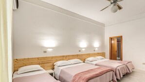 Une chambre d’hôtel avec trois lits, une tête de lit en bois, des murs blancs et un ventilateur de plafond.