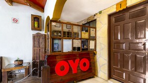 Un mostrador de recepción de madera con un gran letrero 'OYO', un cartel de no fumar, una mesita con una planta en maceta y una puerta marrón.