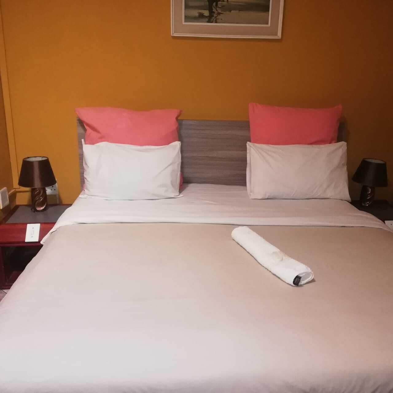 Una cama bien hecha con almohadas blancas y rosadas, una toalla blanca enrollada y un cuadro enmarcado encima.