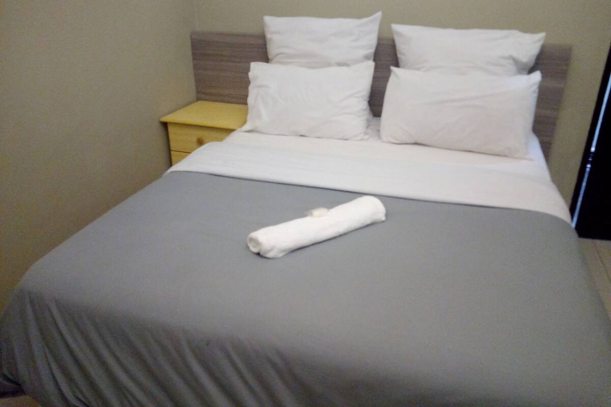 A neatly made bed with white pillows and a folded towel, a wooden headboard, and a small bedside table.