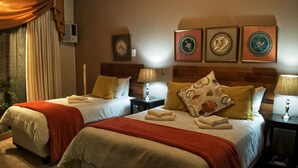 Una habitación de hotel con dos camas, cada una con una colcha rojo y blanco, una mesita de noche con una lámpara y obras de arte enmarcadas en la pared.