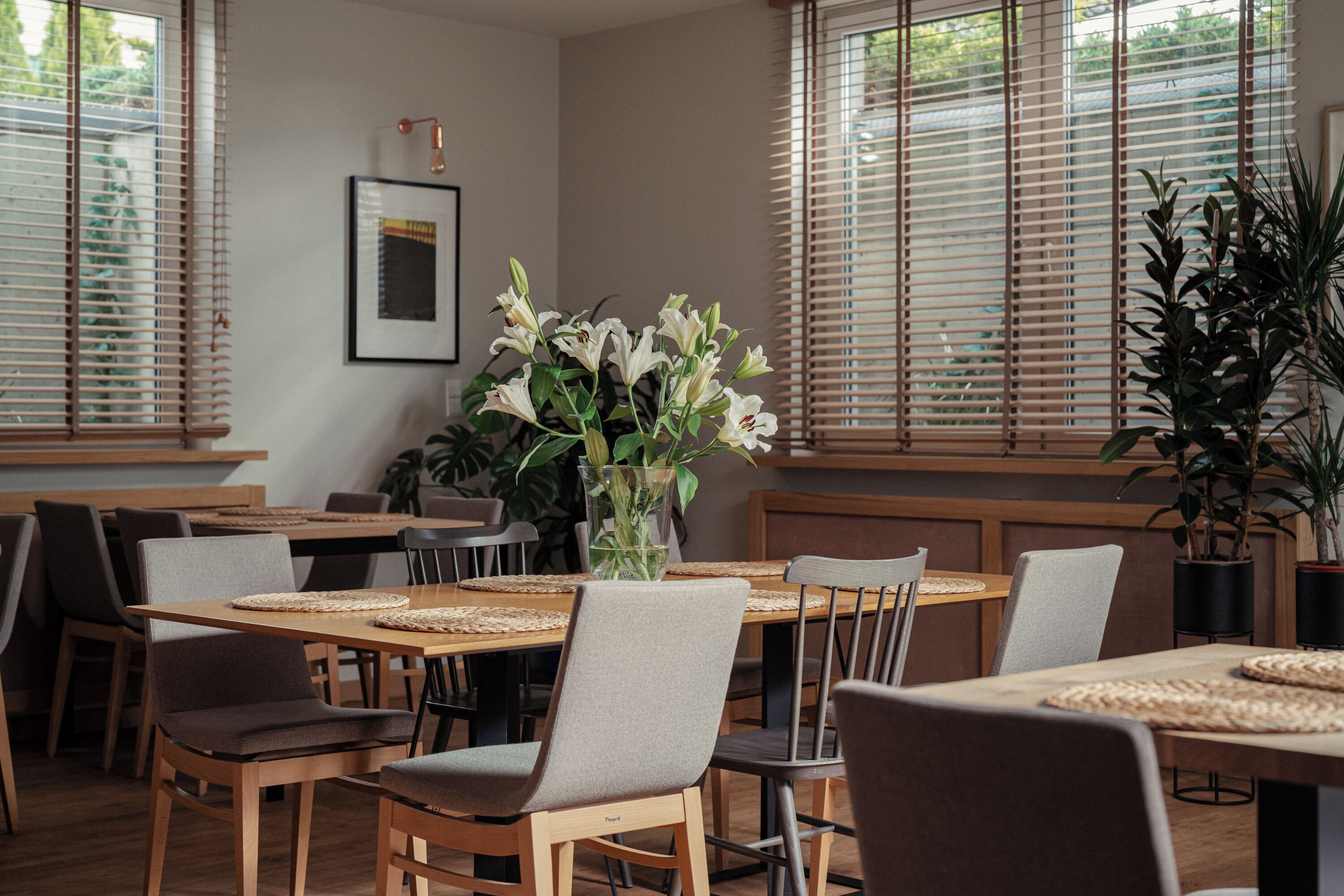 A dining area with wooden tables and chairs, a vase with white flowers, and large windows with blinds.
