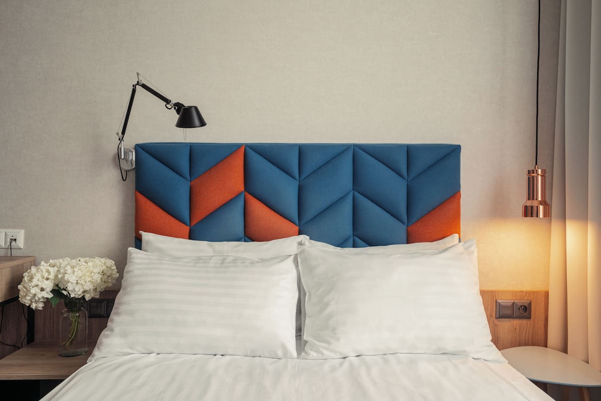 Una cama bien tendida con sábanas blancas, un cabecero moderno con motivos geométricos, una mesita de noche con un jarrón de flores y una lámpara.