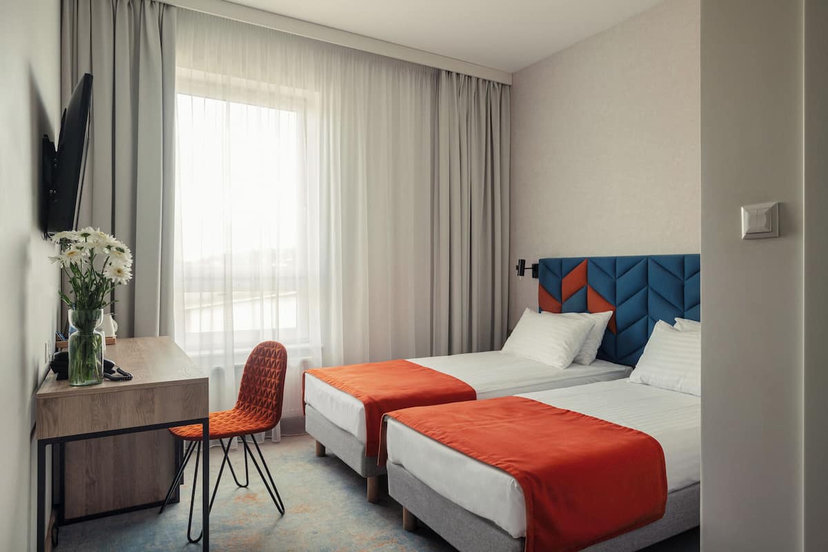 Habitación de hotel con dos camas individuales, un escritorio con silla, un televisor de pantalla plana y un florero con flores.