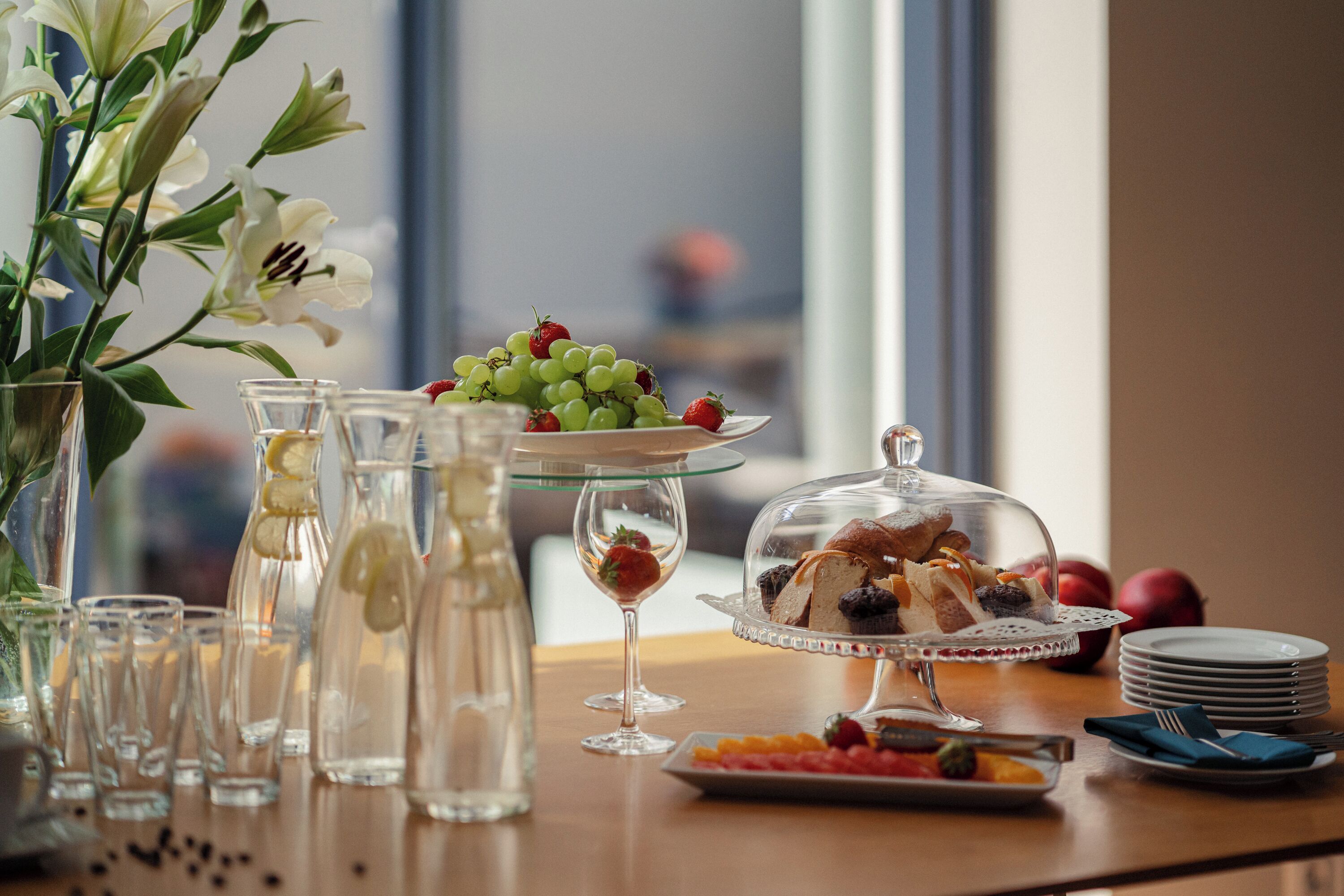 Un bufé de desayuno con diversos alimentos, como uvas, fresas y pasteles, dispuesto en una mesa con cristalería transparente y un jarrón de flores.