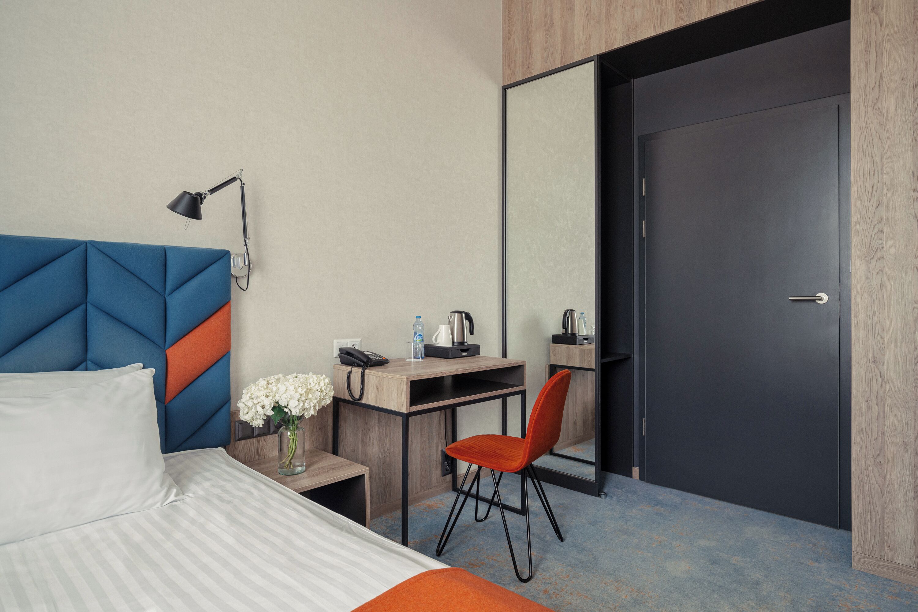 A hotel room with a bed, a desk, a chair, a nightstand, a lamp, a mirror, and a door.
