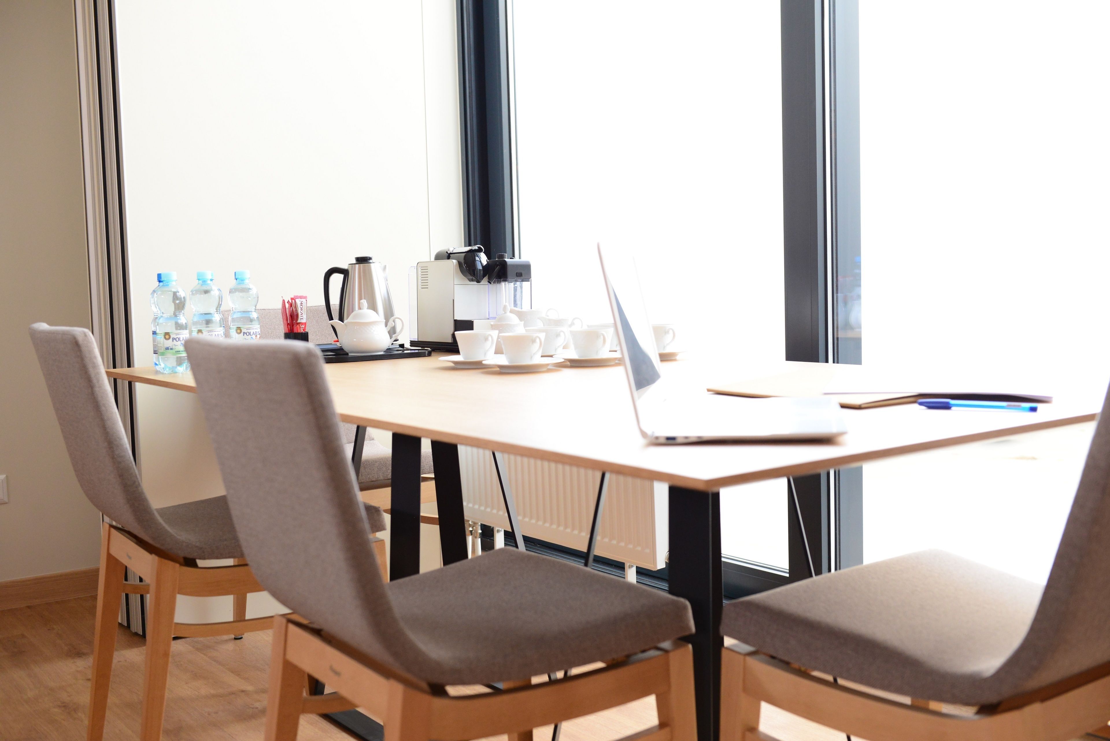 A conference room with a wooden table, two grey cushioned chairs, a laptop, a coffee maker, and water bottles.
