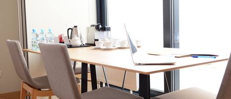 Una sala de conferencias con una mesa de madera, dos sillas grises con cojines, una computadora portátil, una cafetera y botellas de agua.