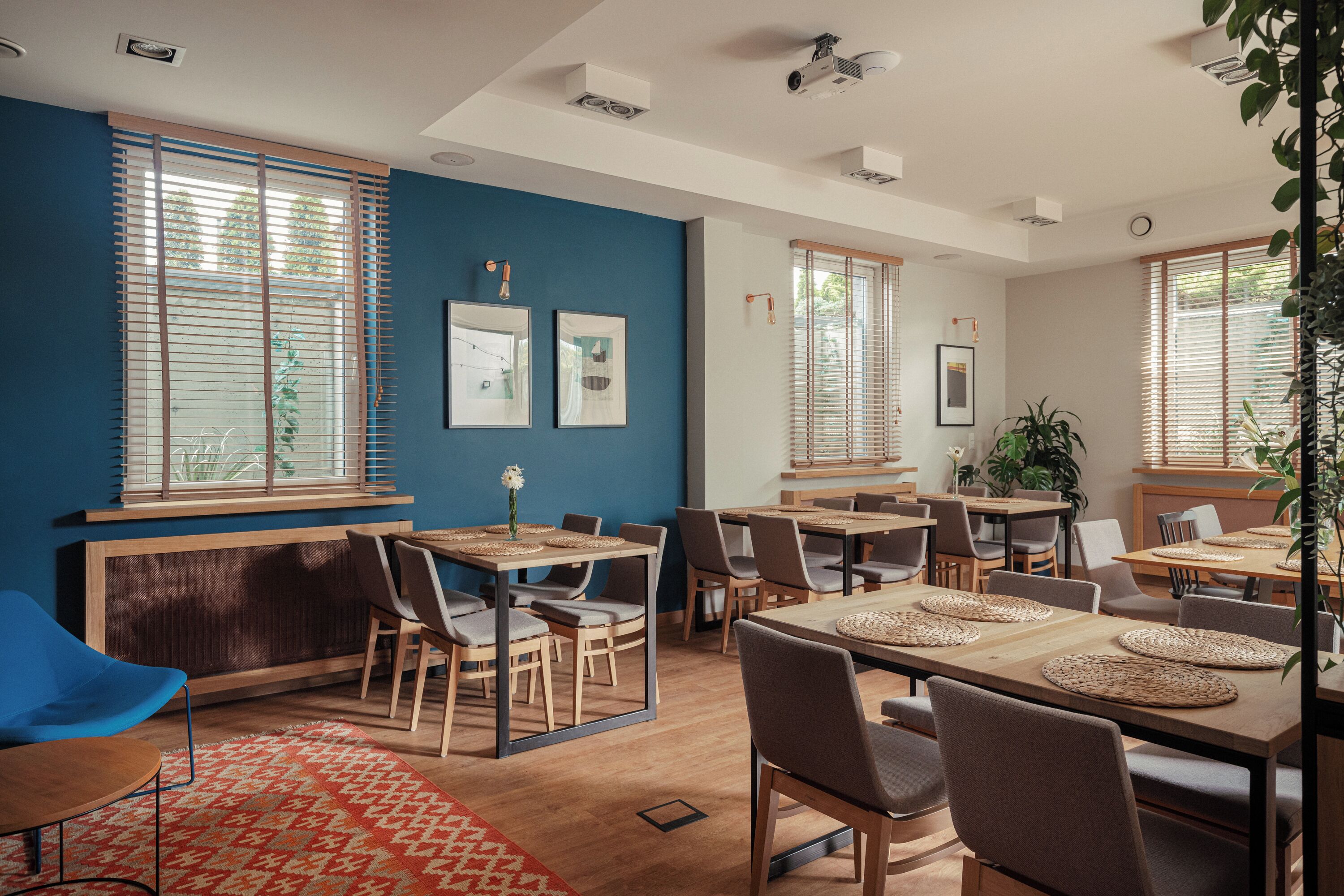 A modern dining area with multiple tables and chairs, large windows with blinds, and a blue accent wall.