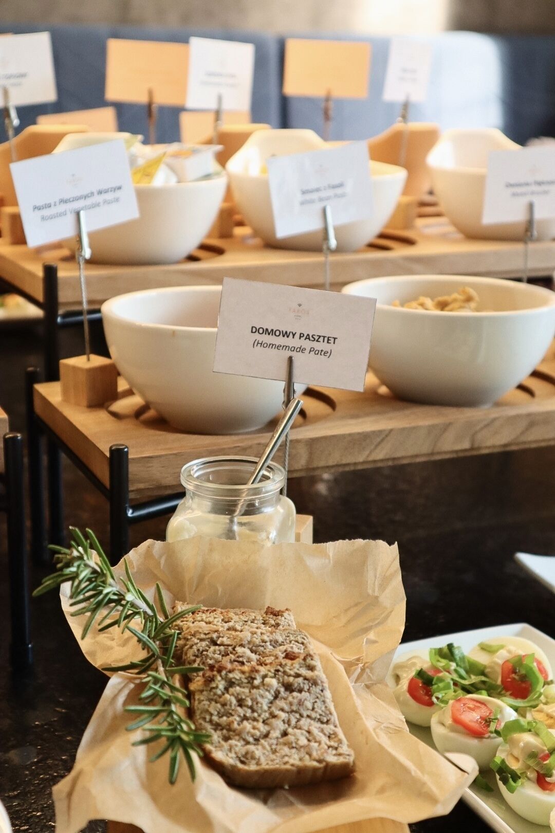 A buffet setup with labeled bowls of food, including a homemade pate, and a jar of milk with a sprig of rosemary.