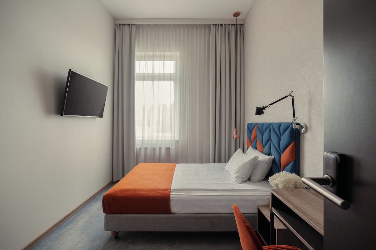 Una habitación de hotel moderna con una cama, un escritorio, una silla, un televisor montado en la pared y una ventana con cortinas.