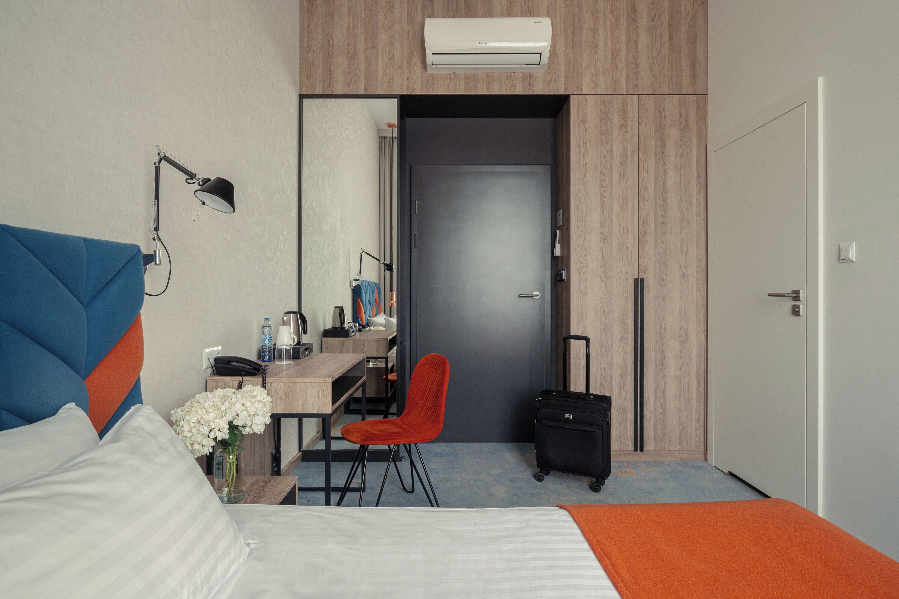 A hotel room with a bed, desk, chair, and a suitcase.