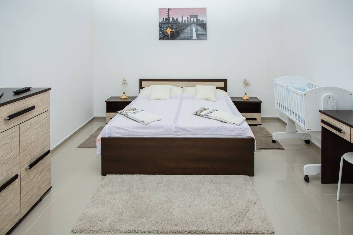 A bedroom with a bed, bedside tables, a crib, a desk, and a cityscape painting on the wall.