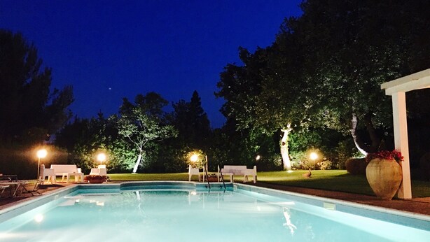 Ein Schwimmbad mit Liegestühlen und einer überdachten Sitzgruppe, umgeben von Bäumen und beleuchtet durch Nachtlichter.