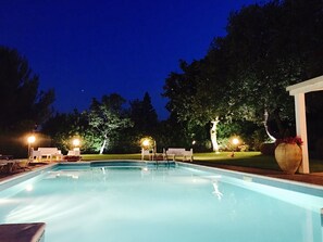 Une piscine dotée de chaises longues et d’un espace salon couvert, entourée d’arbres et illuminée par des jeux de lumière la nuit.