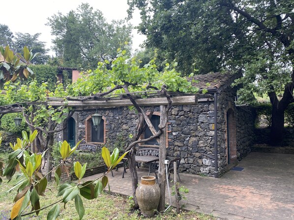 Un edificio de piedra rústico con una pérgola de madera, un jarrón grande y un banco al fondo.