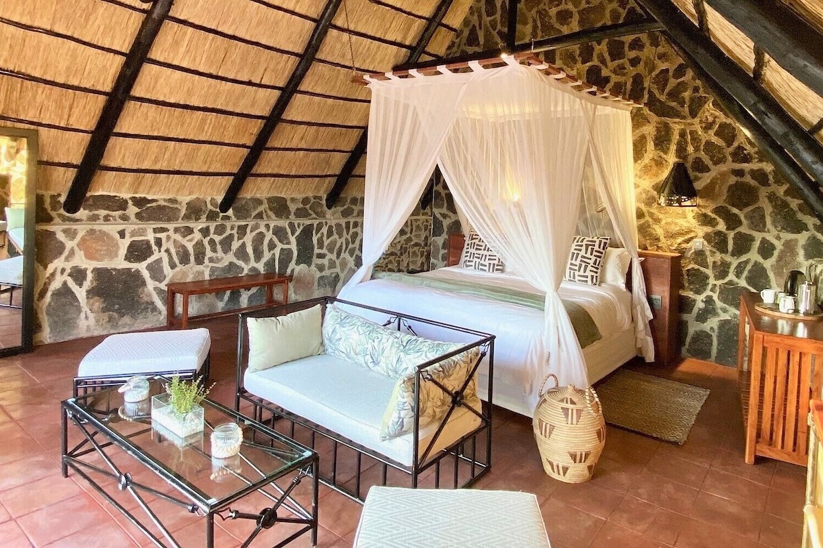 A cozy room with a bed under a canopy, a glass table with a plant, and a stone wall.