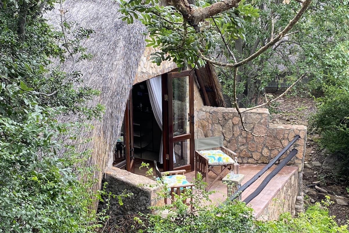 A thatched-roof structure with a glass door, stone walls, and a small outdoor seating area with a table and chairs.