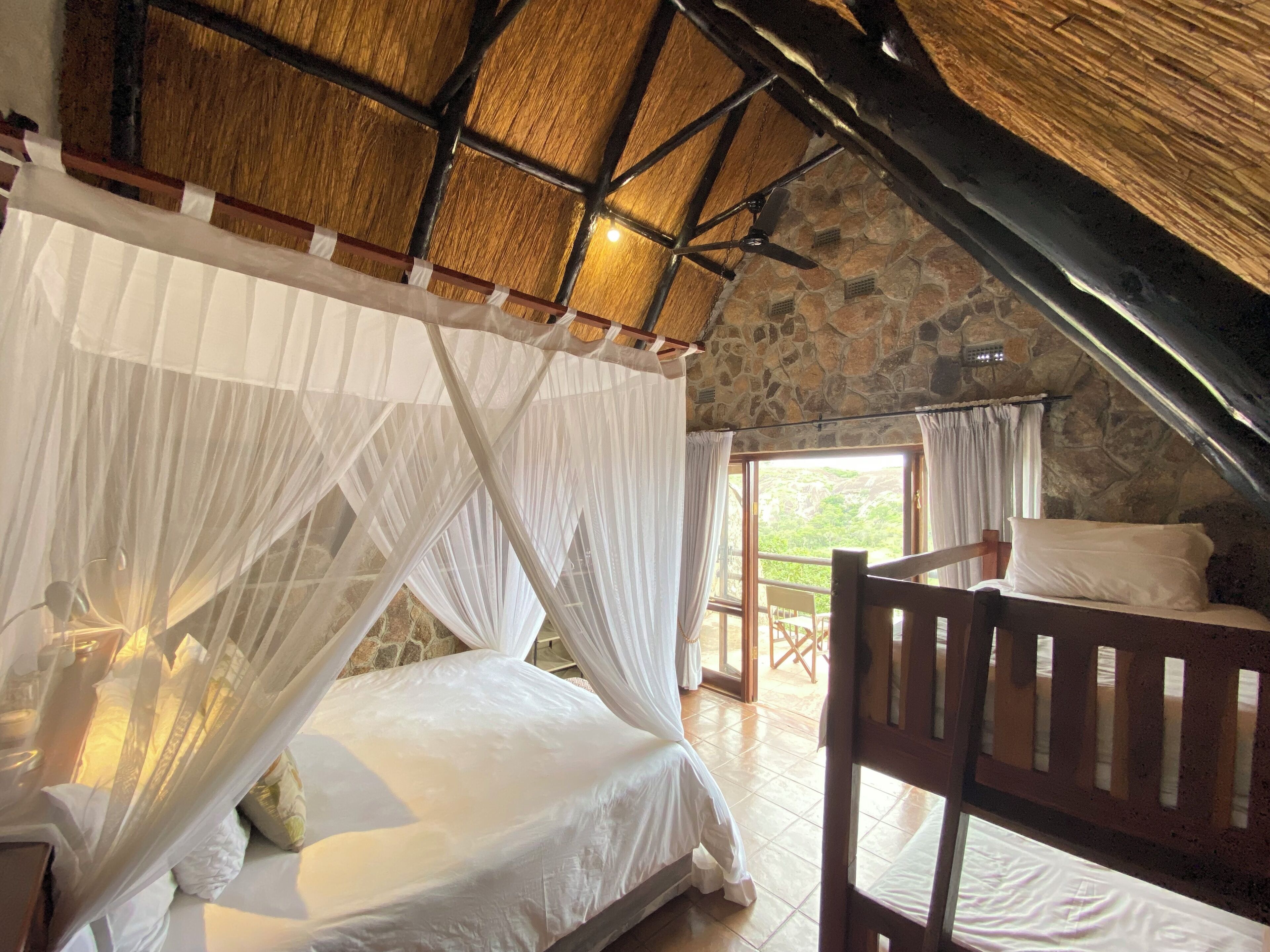 A bedroom with a stone ceiling, a bed with white bedding, a wooden chair, and a view of greenery outside.