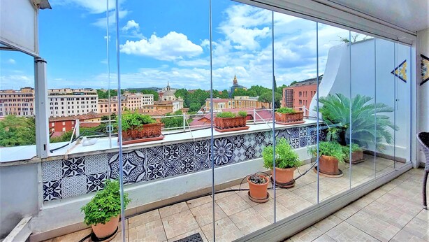 Varanda com vasos de plantas, parede de azulejos e vista para edifícios e uma torre.