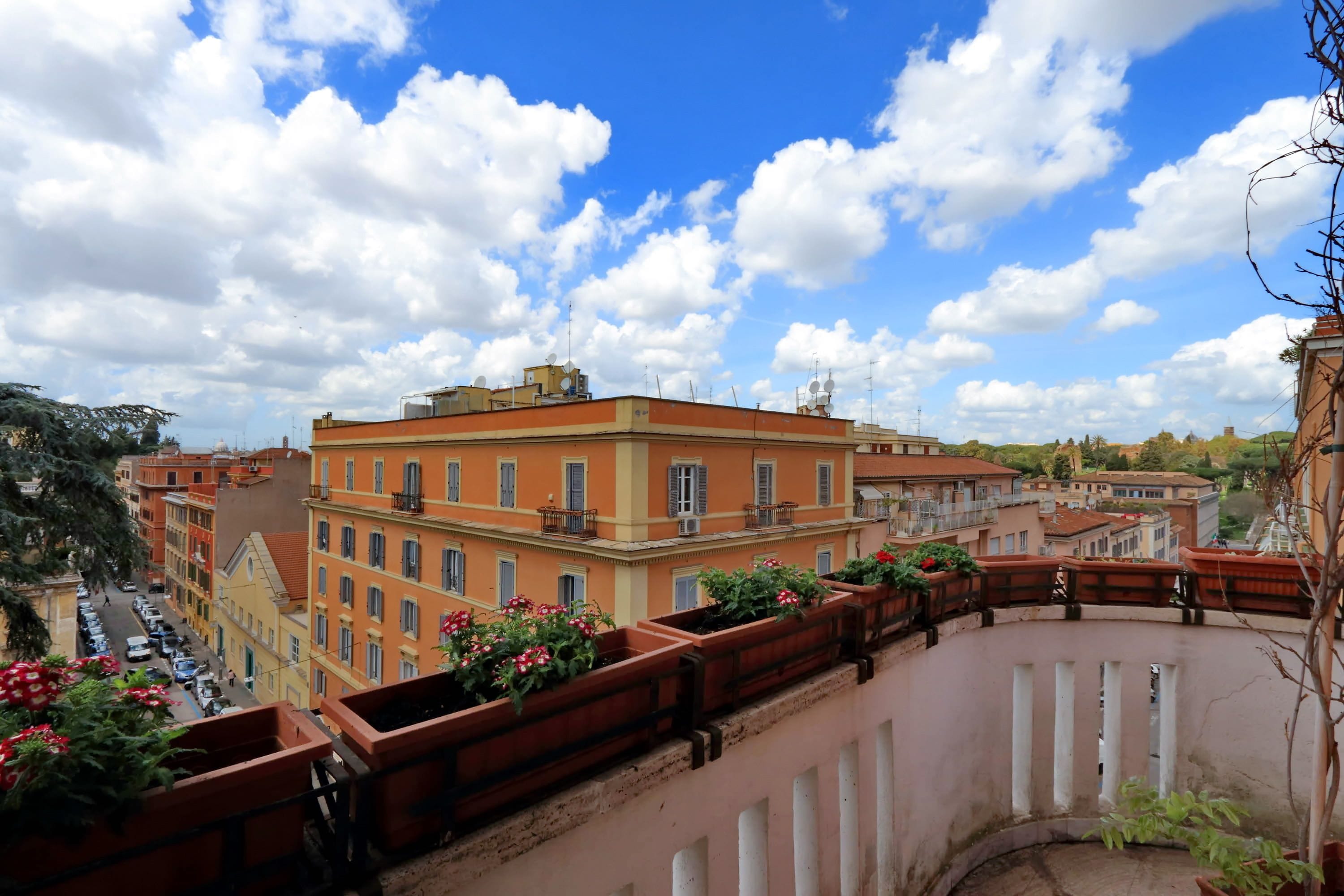 Blick auf eine Stadtlandschaft mit Gebäuden, blauem Himmel und einem geschwungenen Balkon mit Topfpflanzen.