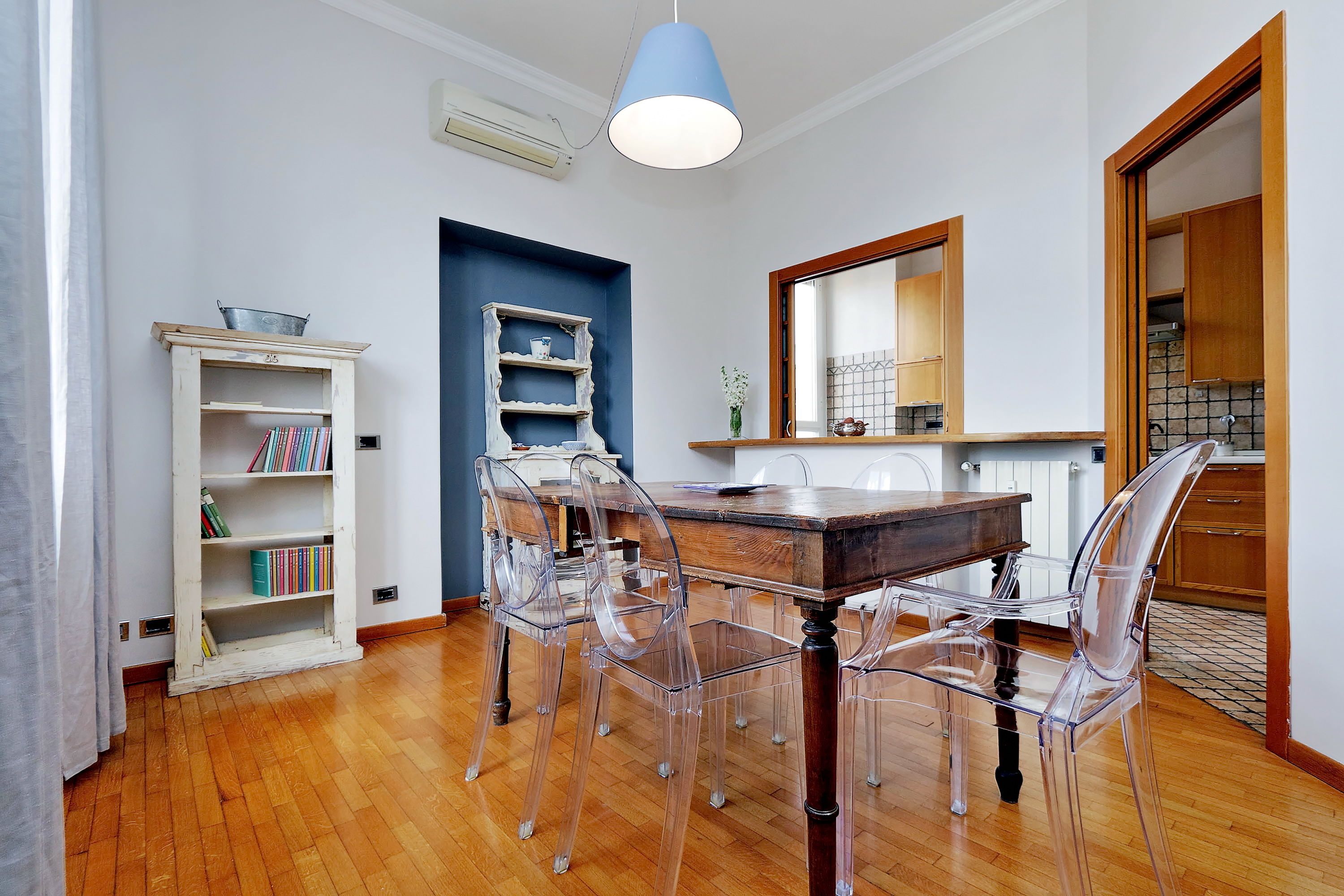 A dining area with a wooden table, clear chairs, a white bookshelf, and a blue pendant light.