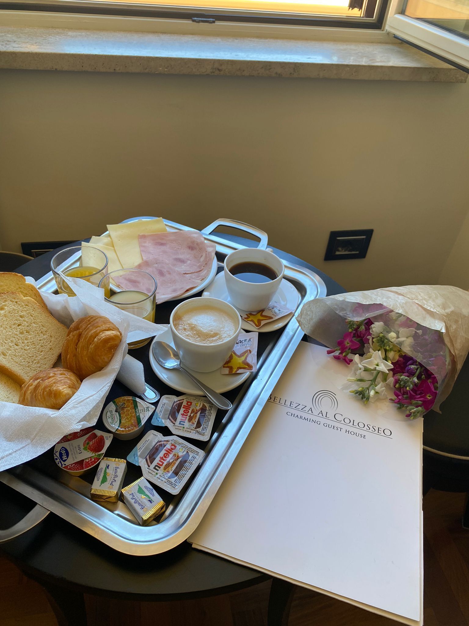 A tray with breakfast items including croissants, cheese, ham, coffee, and pastries, along with a bouquet of flowers and a menu from Settezzo Charming Guest House.