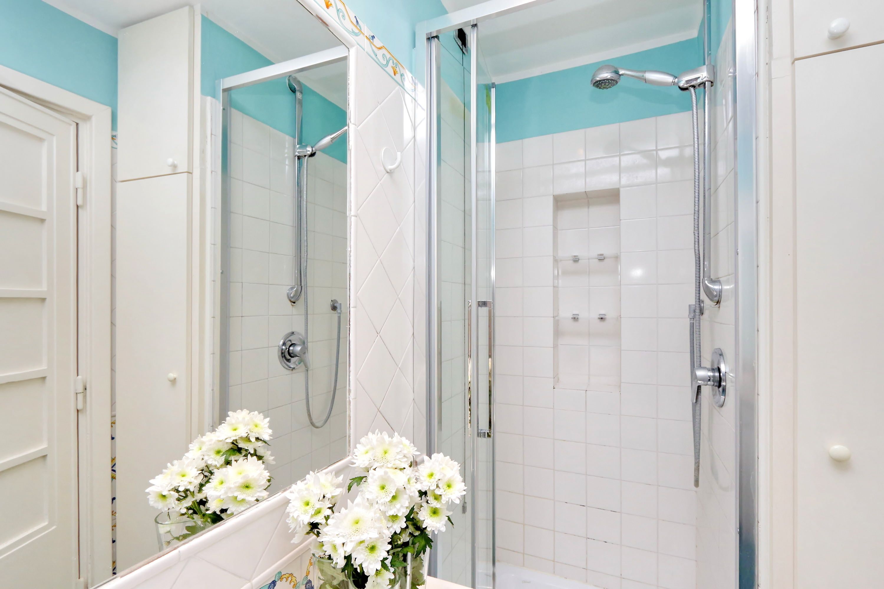 Un baño con ducha de vidrio, azulejos blancos y un espejo con un florero de flores blancas.