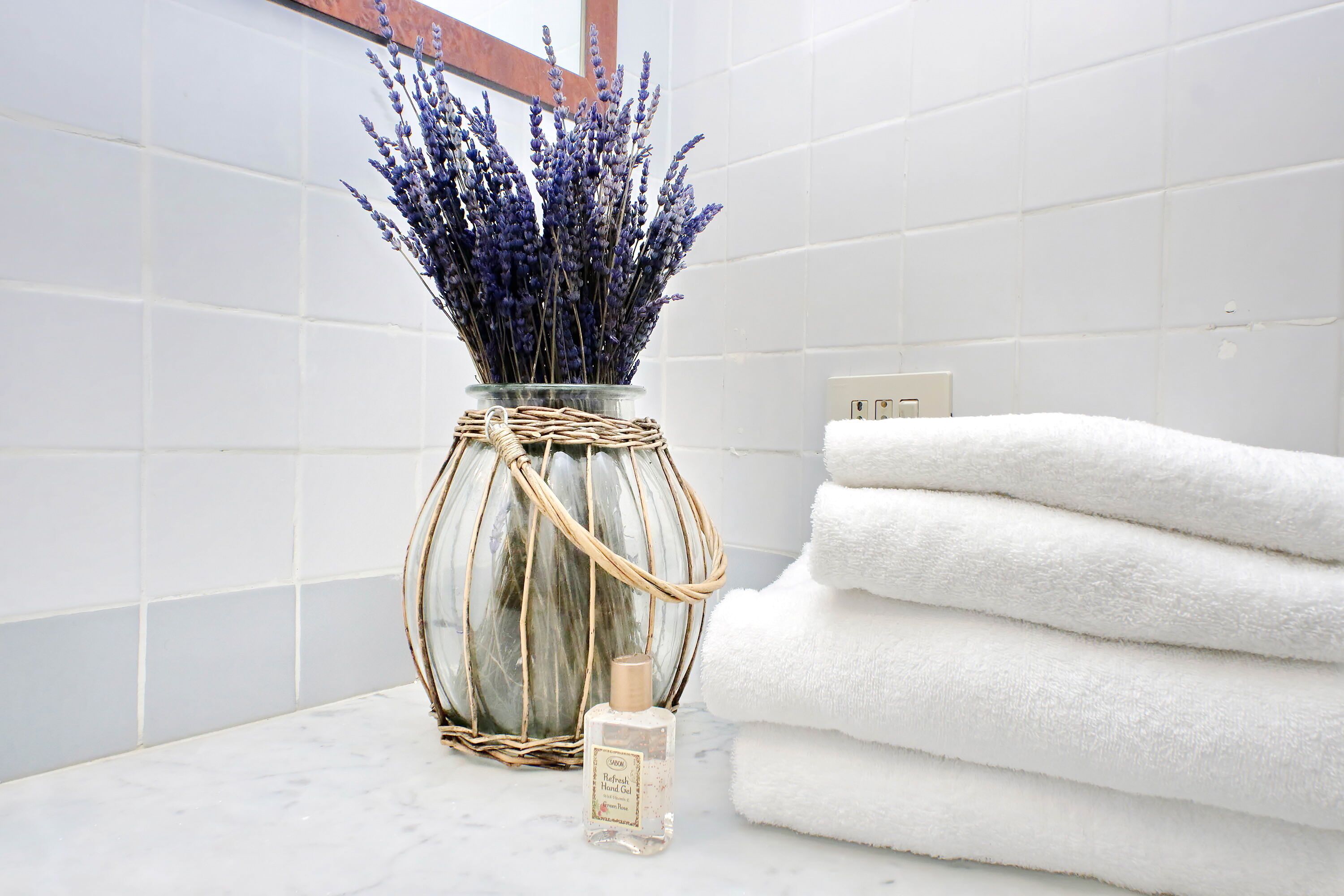 Un baño con azulejos blancos, un florero de vidrio con lavanda, una botella de jabón de manos y toallas blancas apiladas.