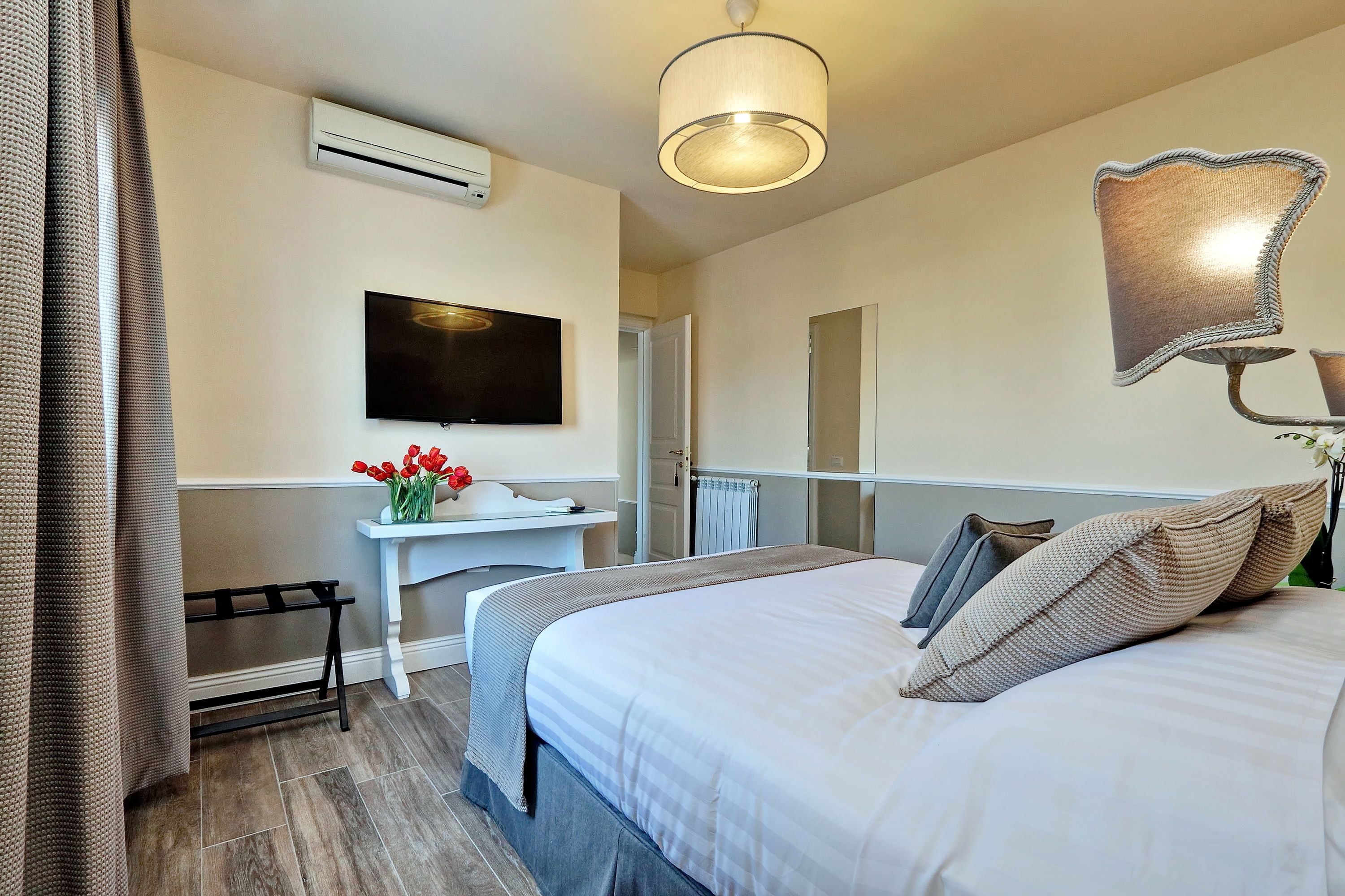 A hotel room with a bed, a desk, a flat-screen TV, an air conditioning unit, and a lamp.
