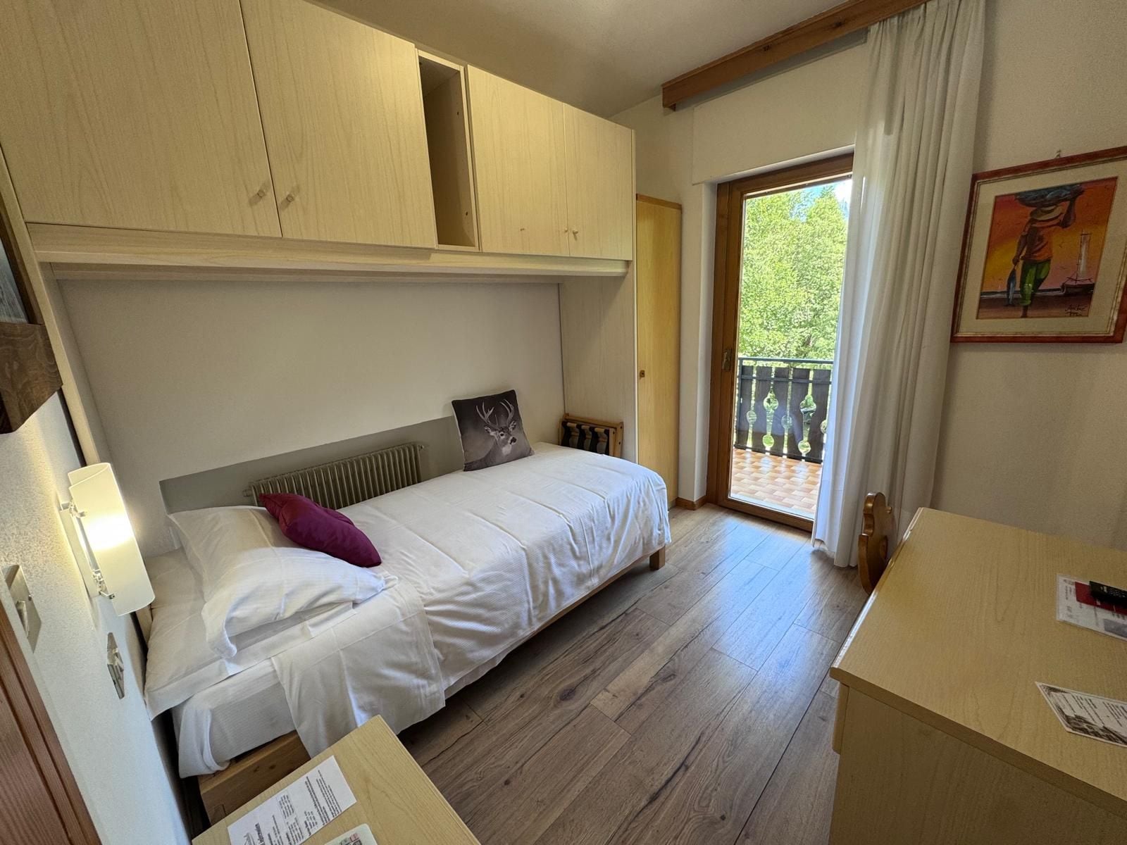 A bedroom with a bed, a nightstand, a wooden wardrobe, a window with a view of greenery, and a painting on the wall.