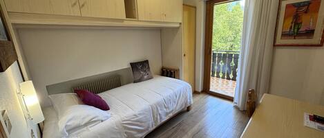 A bedroom with a bed, a nightstand, a wooden wardrobe, a window with a view of greenery, and a painting on the wall.
