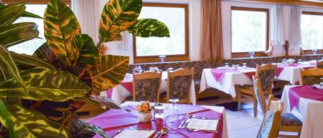 Un restaurante con mesas dispuestas para comer, amplias ventanas y plantas en macetas.